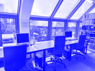 Zu sehen sind drei Arbeitsplätze in der Bibliothek vor einer Fensterfront mit blauen Bürostühlen. Zwei der Arbeitsplätze haben einen Desktop-PC, einer eine Tischlampe. Rechts am Bildrand ist ein Bücherregal zu sehen. Durch das Fenster sieht man einige Pflanzen auf der Dachterasse.
