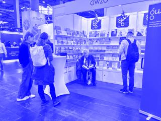 Messestand mit Büchern und Flyern, eine sitzende Person liest, mehrere Personen stehen davor.