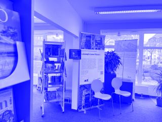 Zwei grüne Stühle vor einem Informationsdisplay mit Text und Bildern in einem hellen Raum mit großen Fenstern und Bücherregalen.
