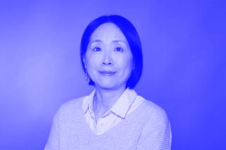 Person mit dunklen, schulterlangen Haaren trägt weißen Hemdkragen unter hellgrauem Pullover, vor einfarbigem Hintergrund.