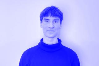 Person mit kurzem Haar trägt einen schwarzen Rollkragenpullover vor weißer Wand.