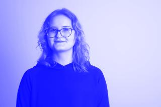 Person mit schulterlangen gewellten Haaren und Brille lächelt leicht in die Kamera und trägt einen dunklen Pullover.