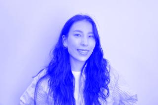 Person mit langen, welligen Haaren trägt gestreiftes Hemd über weißem T-Shirt und Ohrringe vor weißem Hintergrund