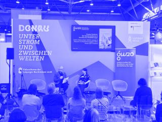 Zwei Personen sitzen auf Stühlen vor einer farbigen Messestandwand mit Texten und Logos, davor Publikum auf transparenten Stühlen in einer Messehalle.