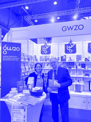 Zwei Personen stehen an einem Messestand des Leibniz-Instituts für Geschichte und Kultur des östlichen Europa mit Büchern und Flyern auf einem Tisch und Regalen im Hintergrund, darüber ein Schild mit der Aufschrift 'GWZO'