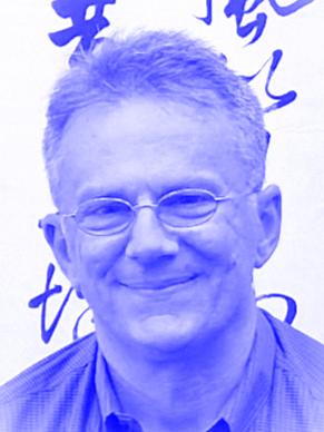 Porträt eines lächelnden Mannes mit Brille und grauem Haar vor einer Wand mit Schrift.