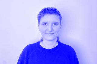 Person mit kurzem Haar trägt einen dunklen Pullover vor weißem Hintergrund.