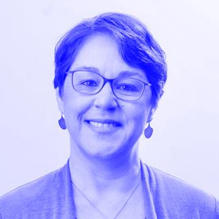 Portrait Manuela Boatca: Person mit kurzen braunen Haaren, grauer Strickjacke und silbernen Ohrringen und Halskette vor hellem Hintergrund.