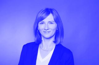 Frau mit schulterlangem, glattem Haar trägt ein schwarzes Sakko und ein weißes V-Ausschnitt-Oberteil vor einfarbigem Hintergrund.