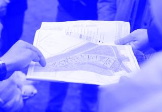 Mehrere Personen betrachten einen farbigen Lageplan mit Gebäuden und Grünflächen, eine Hand zeigt auf den Plan.