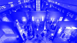 Blick auf die Ausstellung »Kaiser Karl IV. 1316-2016« in der Wallenstein Reithalle, Nationalgalerie Prag. Mehrere Vitrinen mit Kunstwerken, Besucher und zwei ausgestellte, beleuchtete Statuen im Zentrum.