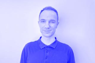 Person mit kurzen Haaren trägt ein dunkelgraues Poloshirt mit Knopfleiste vor weißer Wand.