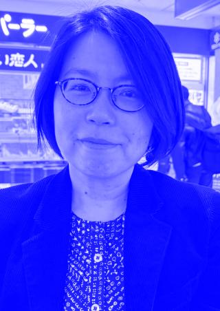 Frau mit schwarzem kurzem Haar, Brille und schwarzem Sakko vor einem Laden mit japanischer Beschilderung.