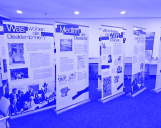 Fünf aufstellbare Roll-ups mit Text und historischen Schwarz-weiß-Fotos zur Dissidenz stehen in einem Raum auf grauem Teppichboden.