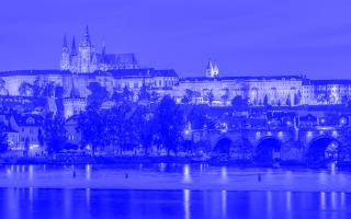 Abendliche Ansicht der Prager Burg und der Karlsbrücke in Prag, Tschechien. Die historischen Gebäude sind golden beleuchtet und spiegeln sich im ruhigen Wasser der Moldau. Die Brücke mit ihren Steinbögen und Statuen verbindet die beiden Ufer der Stadt. Der Himmel zeigt ein tiefes Blau der Dämmerung.