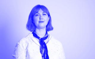 Porträt einer Person mit schulterlangen Haaren, die eine weiße Bluse und ein dunkles Halstuch trägt.