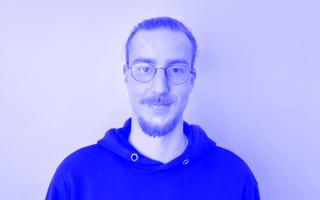 Mann mit zusammengebundenem Haar und Bart trägt schwarzen Kapuzenpullover vor weißem Hintergrund.