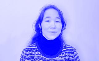 Person mit langen, gewellten Haaren trägt einen gemusterten Pullover über einem schwarzen Rollkragenpullover vor einfarbigem Hintergrund.