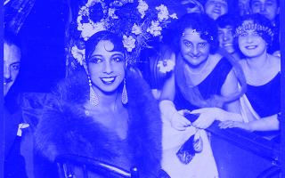 Josephine Baker sitzt elegant in einem Stuhl, trägt ein aufwändiges Kopfschmuck mit Blumen, ein langes Kleid und einen Pelz, umgeben von mehreren Personen in Abendkleidung.