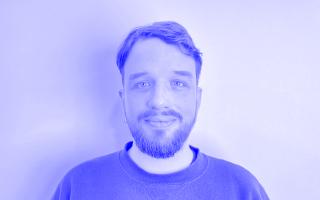 Person mit kurzem Haar und Bart trägt ein Sweatshirt vor einfarbigem Hintergrund