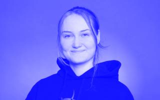 Person mit zusammengebundenem Haar trägt schwarzen Kapuzenpullover vor einfarbigem Hintergrund.