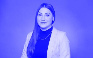 Person mit langen glatten Haaren, schwarzem Rollkragenpullover, heller Jacke und Perlenkette vor einfarbigem Hintergrund.