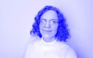 Person mit lockigem, schulterlangem Haar trägt einen weißen Rollkragenpullover vor hellem Hintergrund.