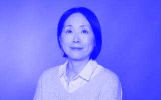 Person mit dunklen, schulterlangen Haaren trägt weißen Hemdkragen unter hellgrauem Pullover, vor einfarbigem Hintergrund.