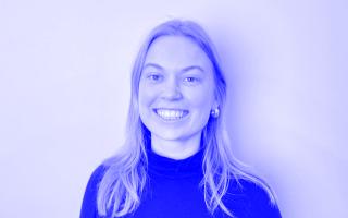 Person mit langen Haaren, die einen schwarzen Rollkragenpullover trägt, vor weißem Hintergrund.