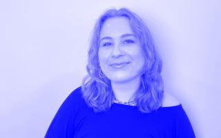 Person mit schulterlangen gewellten Haaren trägt ein schwarzes Oberteil mit Ausschnitt und eine Halskette vor hellem Hintergrund