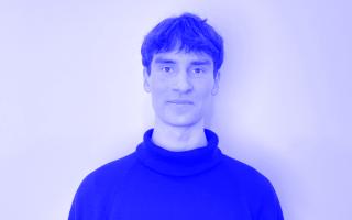 Person mit kurzem Haar trägt einen schwarzen Rollkragenpullover vor weißer Wand.