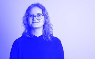Person mit schulterlangen gewellten Haaren und Brille lächelt leicht in die Kamera und trägt einen dunklen Pullover.