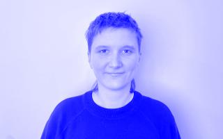 Person mit kurzem Haar trägt einen dunklen Pullover vor weißem Hintergrund.