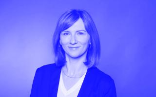 Frau mit schulterlangem, glattem Haar trägt ein schwarzes Sakko und ein weißes V-Ausschnitt-Oberteil vor einfarbigem Hintergrund.