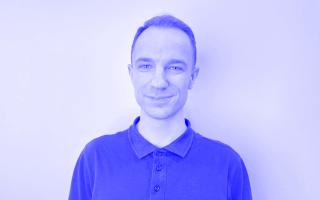 Person mit kurzen Haaren trägt ein dunkelgraues Poloshirt mit Knopfleiste vor weißer Wand.