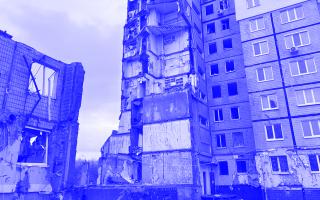 Zerstörtes mehrstöckiges Wohngebäude mit freigelegten Innenwänden und Trümmern, daneben teilweise eingestürztes Gebäude, bewölkter Himmel.