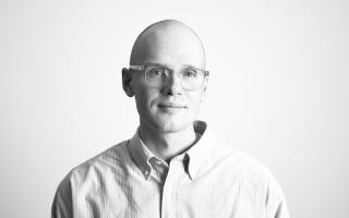 Porträt einer Person mit Brille und Hemd vor hellem Hintergrund.