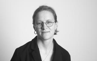 Schwarz-weiße Porträtaufnahme einer Person mit Brille, Ohrringen und zurückgebundenem Haar, die leicht lächelt.