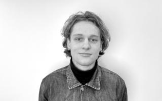 Person trägt Jeansjacke über dunklem Rollkragenpullover vor weißem Hintergrund.