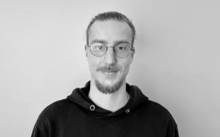 Mann mit zusammengebundenem Haar und Bart trägt schwarzen Kapuzenpullover vor weißem Hintergrund.