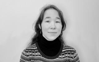 Person mit langen, gewellten Haaren trägt einen gemusterten Pullover über einem schwarzen Rollkragenpullover vor einfarbigem Hintergrund.