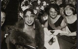 Josephine Baker sitzt elegant in einem Stuhl, trägt ein aufwändiges Kopfschmuck mit Blumen, ein langes Kleid und einen Pelz, umgeben von mehreren Personen in Abendkleidung.
