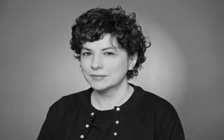 Person mit welligem, kurzem Haar, die ein schwarzes Oberteil mit Knöpfen trägt, vor einfarbigem Hintergrund