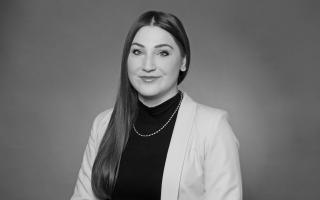 Person mit langen glatten Haaren, schwarzem Rollkragenpullover, heller Jacke und Perlenkette vor einfarbigem Hintergrund.