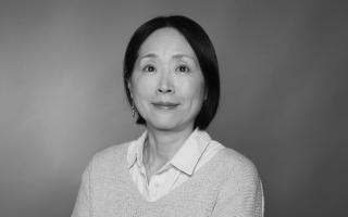 Person mit dunklen, schulterlangen Haaren trägt weißen Hemdkragen unter hellgrauem Pullover, vor einfarbigem Hintergrund.