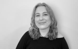 Person mit schulterlangen gewellten Haaren trägt ein schwarzes Oberteil mit Ausschnitt und eine Halskette vor hellem Hintergrund