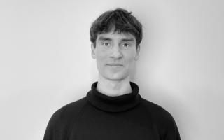 Person mit kurzem Haar trägt einen schwarzen Rollkragenpullover vor weißer Wand.