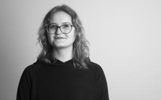 Person mit schulterlangen gewellten Haaren und Brille lächelt leicht in die Kamera und trägt einen dunklen Pullover.