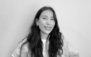 Person mit langen, welligen Haaren trägt gestreiftes Hemd über weißem T-Shirt und Ohrringe vor weißem Hintergrund