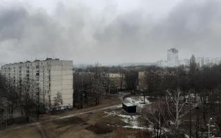 Blick auf eine städtische Landschaft mit grauen Wolken und trübem Himmel. Im Vordergrund sind kahle Bäume und ein leerer Platz sichtbar. Mehrere Wohngebäude sind im Hintergrund zu sehen.